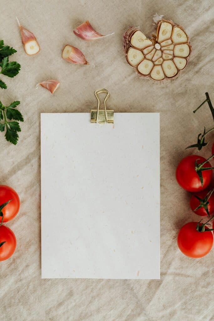 Top view of empty clipboard with golden paper binder placed on linen tablecloth among delicious red tomatoes on branches together with chopped garlic and green parsley suitable for recipe or menu placement